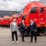 Three businessmen in suits shake hands beside red modern trains outdoors in a rail yard.