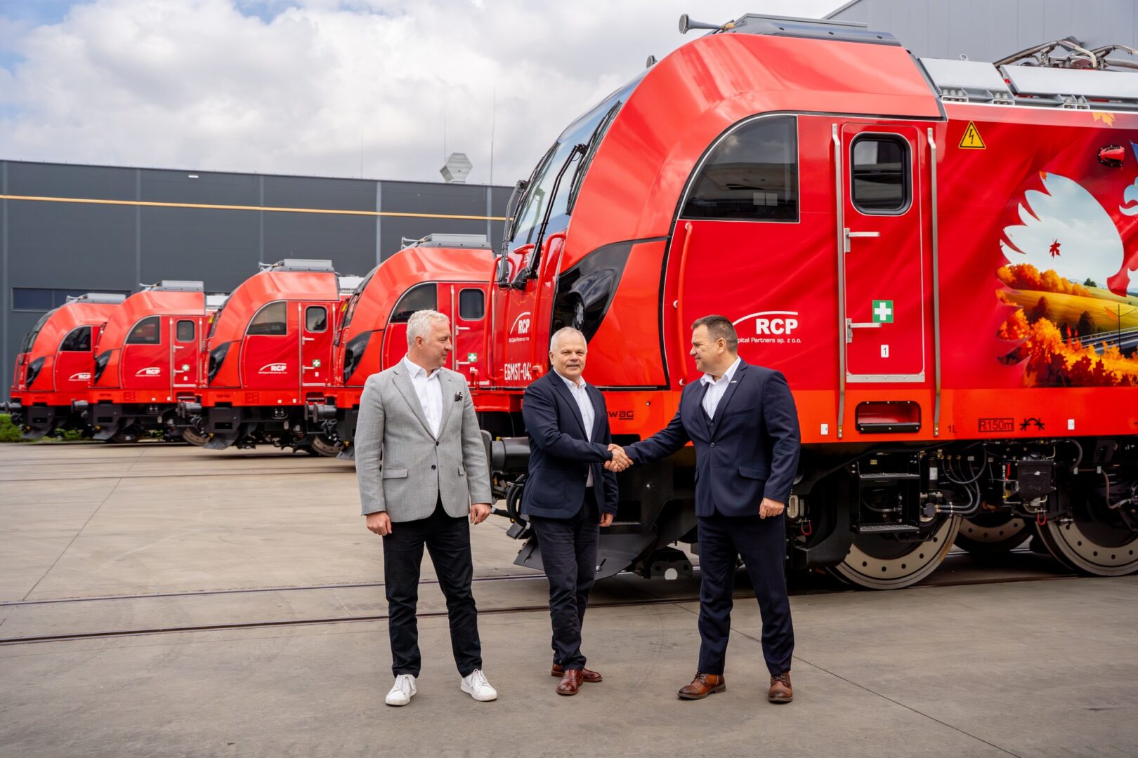 Three businessmen in suits shake hands beside red modern trains outdoors in a rail yard.
