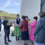Group of people outside a modern building; a woman in folk dress hands items to a line of attendees as others hold flowers and boxes show