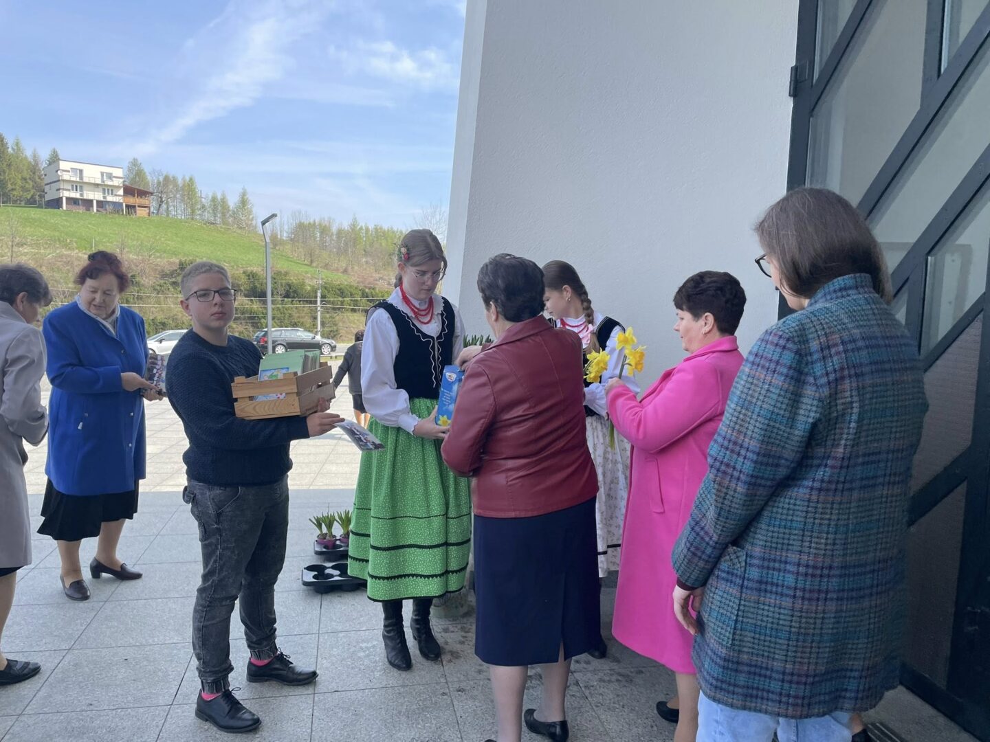 Group of people outside a modern building; a woman in folk dress hands items to a line of attendees as others hold flowers and boxes show