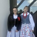 Two women in traditional folk dresses pose together on a balcony, one with a black jacket and red ribbon, the other with a blue floral dress and red bead necklace.