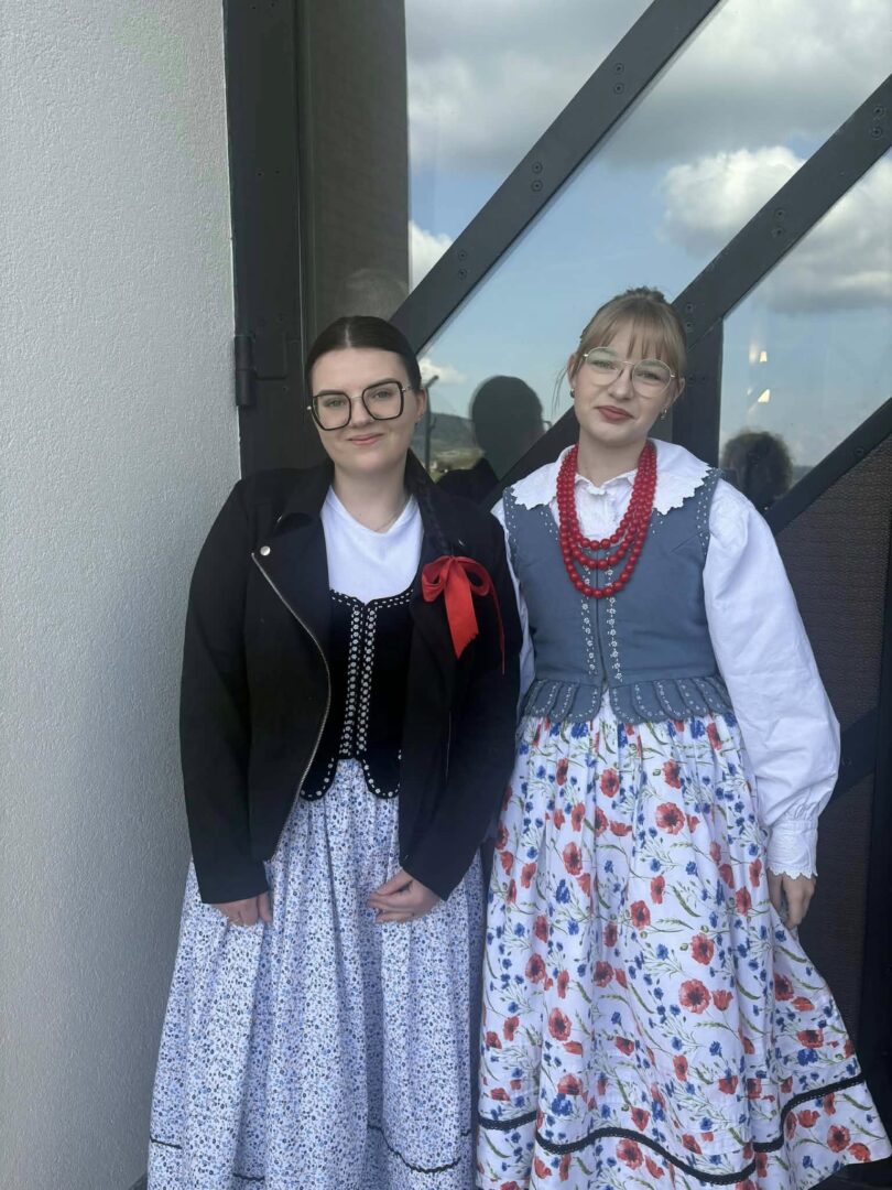 Two women in traditional folk dresses pose together on a balcony, one with a black jacket and red ribbon, the other with a blue floral dress and red bead necklace.