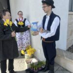 Elderly woman hands yellow daffodils to a man in traditional folk costume; a girl in a floral dress watches, with potted plants and boxes on the ground nearby.