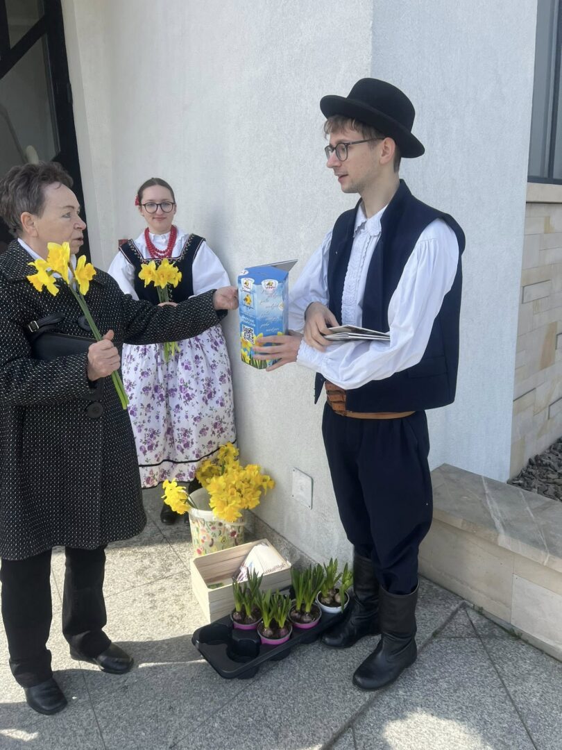 Elderly woman hands yellow daffodils to a man in traditional folk costume; a girl in a floral dress watches, with potted plants and boxes on the ground nearby.