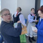 Boy with glasses in a dark blue sweater holding a wooden crate of books, standing near a gray wall as two girls in traditional dresses and an older woman in a blue coat converse nearby outdoors.