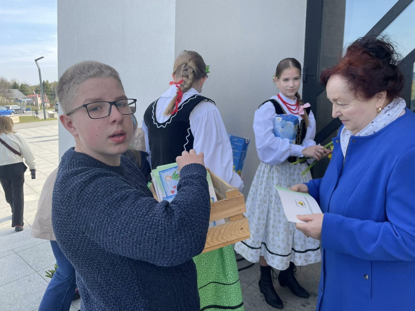 Boy with glasses in a dark blue sweater holding a wooden crate of books, standing near a gray wall as two girls in traditional dresses and an older woman in a blue coat converse nearby outdoors.