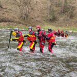 Rescue team in red and yellow suits crossing a river, helmets on, linked for support in flowing water