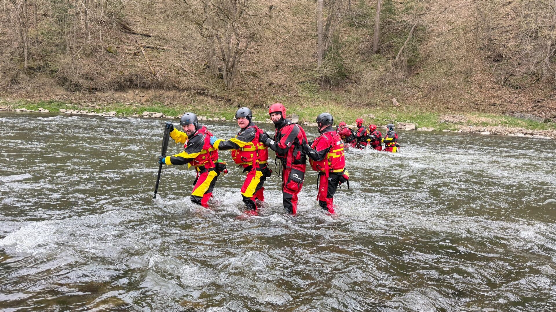 Rescue team in red and yellow suits crossing a river, helmets on, linked for support in flowing water