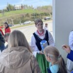 People gathered outdoors near a building; a woman in traditional folk costume holds a blue carton while others watch.