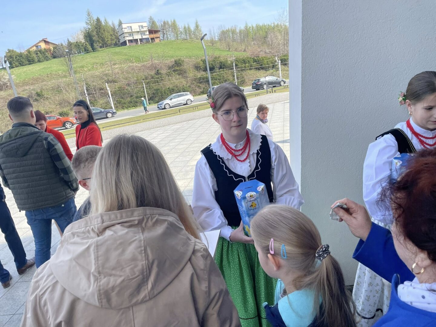 People gathered outdoors near a building; a woman in traditional folk costume holds a blue carton while others watch.