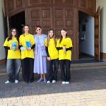 Five people pose in front of a large wooden door; four wear yellow shirts and dark pants, one in a lavender dress with sunglasses, holding books or gifts.