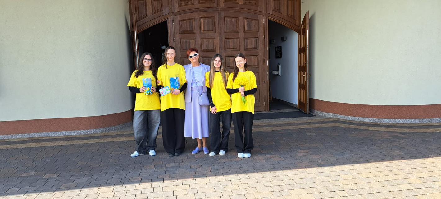 Five people pose in front of a large wooden door; four wear yellow shirts and dark pants, one in a lavender dress with sunglasses, holding books or gifts.