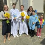 Six people stand outside a stone building, each holding yellow daffodil bouquets, with two in white robes and others in casual clothes.
