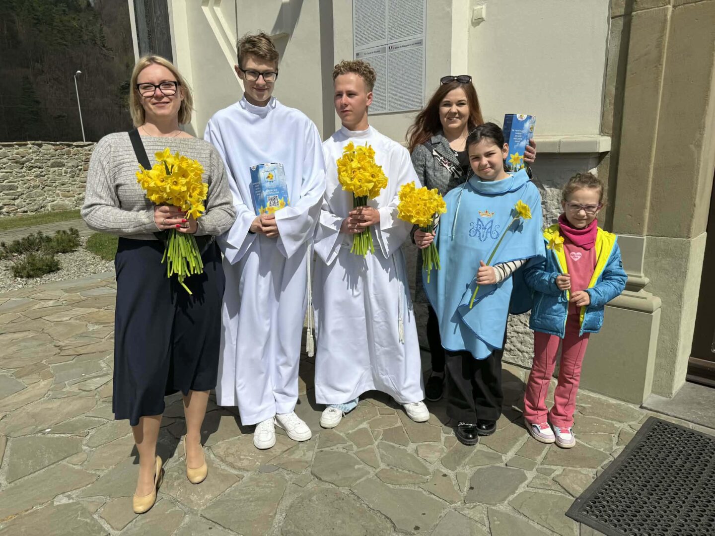Six people stand outside a stone building, each holding yellow daffodil bouquets, with two in white robes and others in casual clothes.