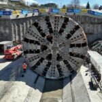 Giant circular tunnel boring machine head in a construction pit, with workers and a red crane nearby.