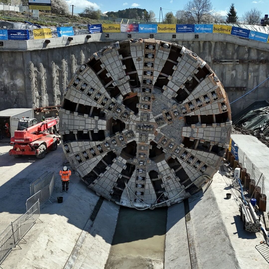 Giant circular tunnel boring machine head in a construction pit, with workers and a red crane nearby.