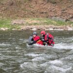 Two people in red life jackets and helmets stand waist-deep in a fast-moving river near a rocky, grassy shore, bracing against the current.