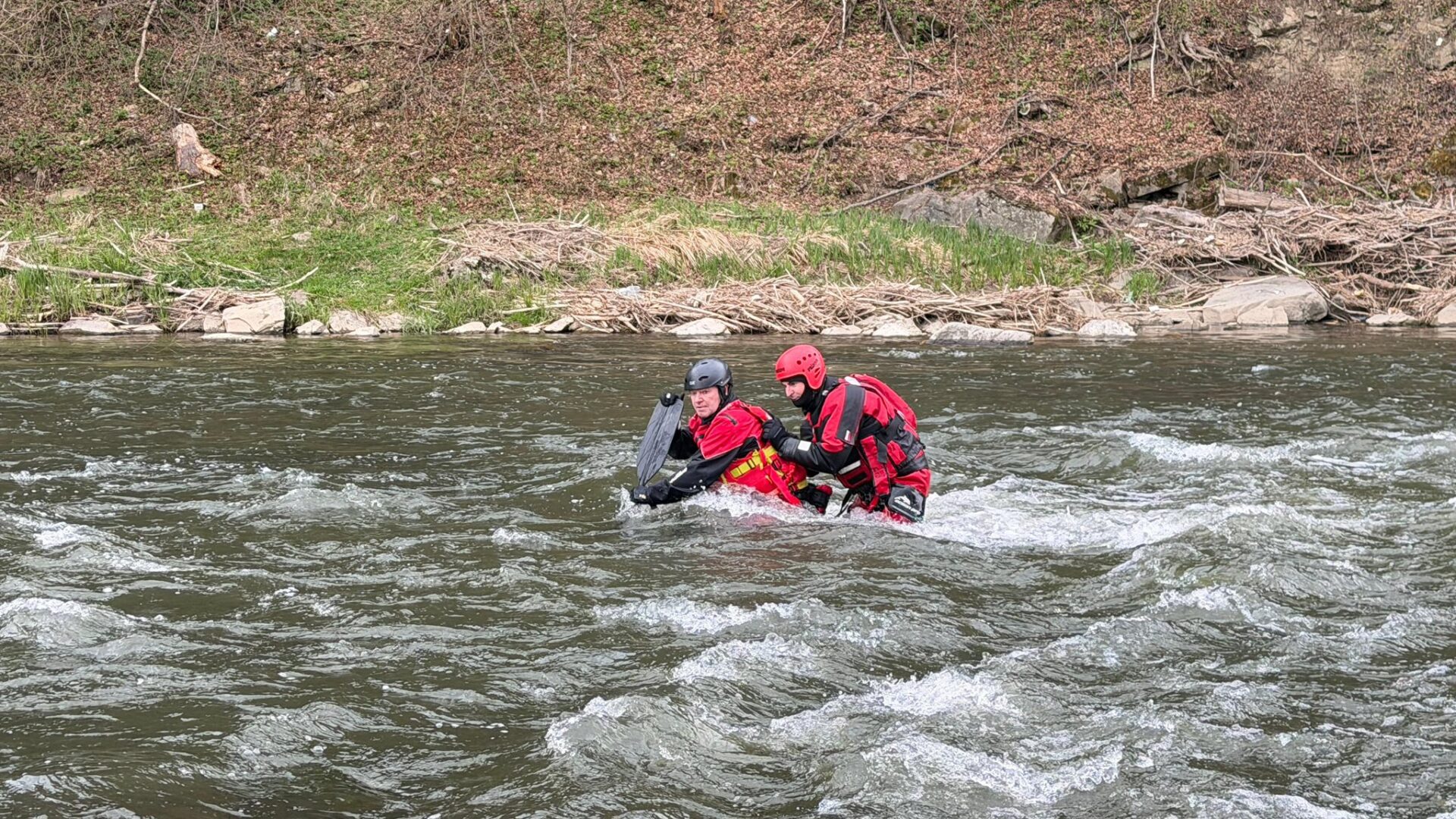 Two people in red life jackets and helmets stand waist-deep in a fast-moving river near a rocky, grassy shore, bracing against the current.