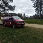 Red emergency vehicle (GOPR) van with blue stripe parked on grass beside a dirt road, trailer attached, trees and houses in the background under a cloudy sky.