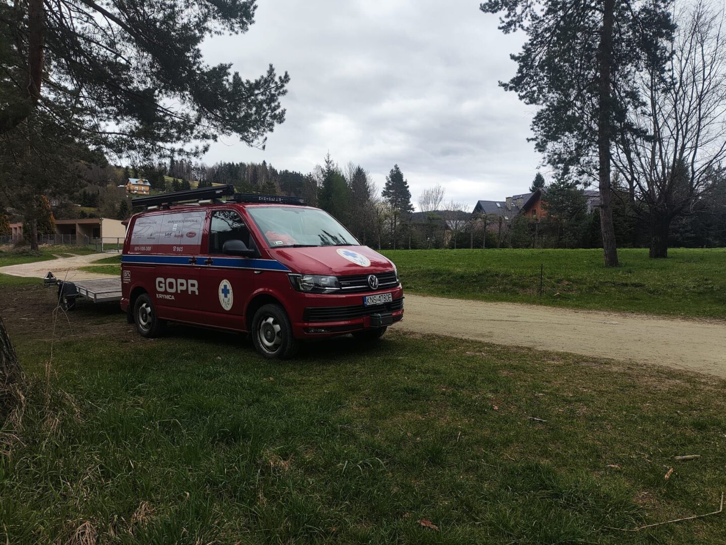 Red emergency vehicle (GOPR) van with blue stripe parked on grass beside a dirt road, trailer attached, trees and houses in the background under a cloudy sky.