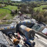 Aerial view of a large construction site with a tunnel boring machine in a deep pit, a tall crane, and heavy equipment surrounded by workers.