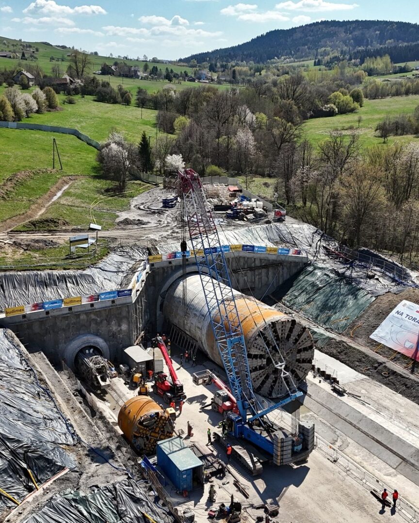 Aerial view of a large construction site with a tunnel boring machine in a deep pit, a tall crane, and heavy equipment surrounded by workers.