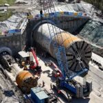 An active construction site with a large tunnel boring machine in a rectangular pit, workers nearby, and equipment set against surrounding hills and trees.