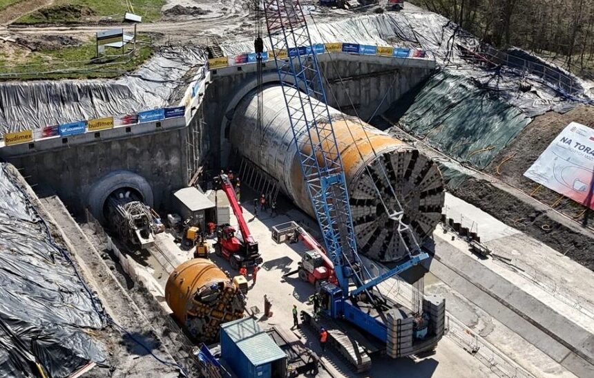 An active construction site with a large tunnel boring machine in a rectangular pit, workers nearby, and equipment set against surrounding hills and trees.