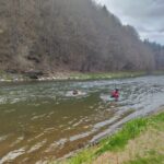 Two people in red life jackets wading in a calm river, with a forested bank and cloudy sky in the background.