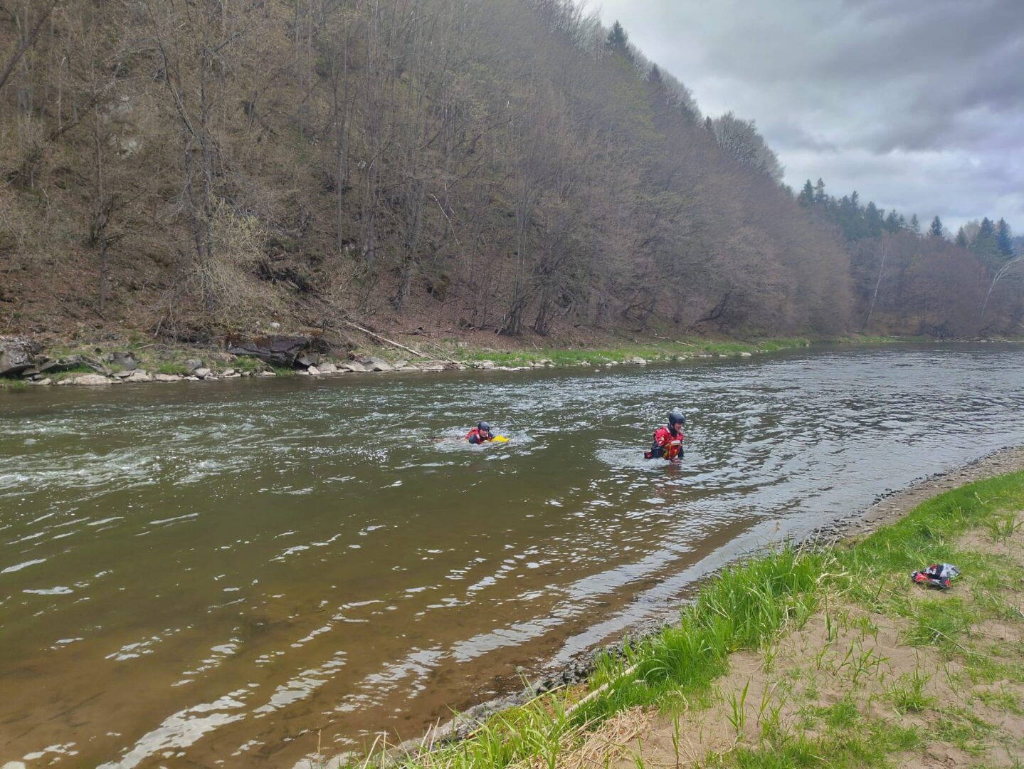 Two people in red life jackets wading in a calm river, with a forested bank and cloudy sky in the background.