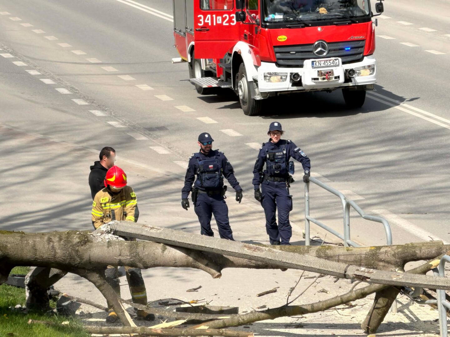 Firefighter in red helmet and two police officers assess a fallen tree blocking a sidewalk, with a red fire truck on the road behind.