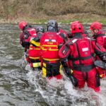 Group of rescue workers in red wetsuits and helmets wading through a river, preparing to reach the far bank.