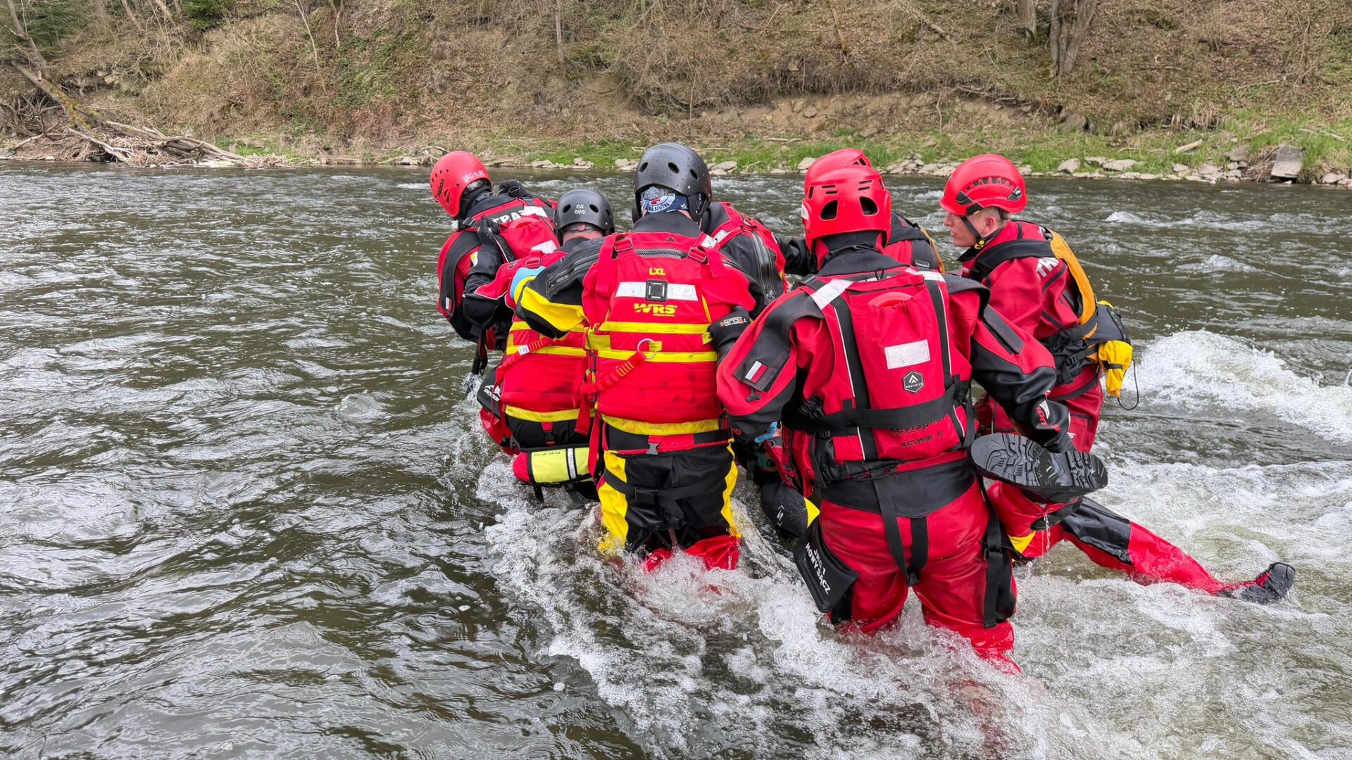 Group of rescue workers in red wetsuits and helmets wading through a river, preparing to reach the far bank.