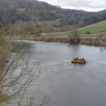 Six people in a yellow inflatable raft with red life jackets float on a wide river with green hills in the background and a tractor on a road nearby.