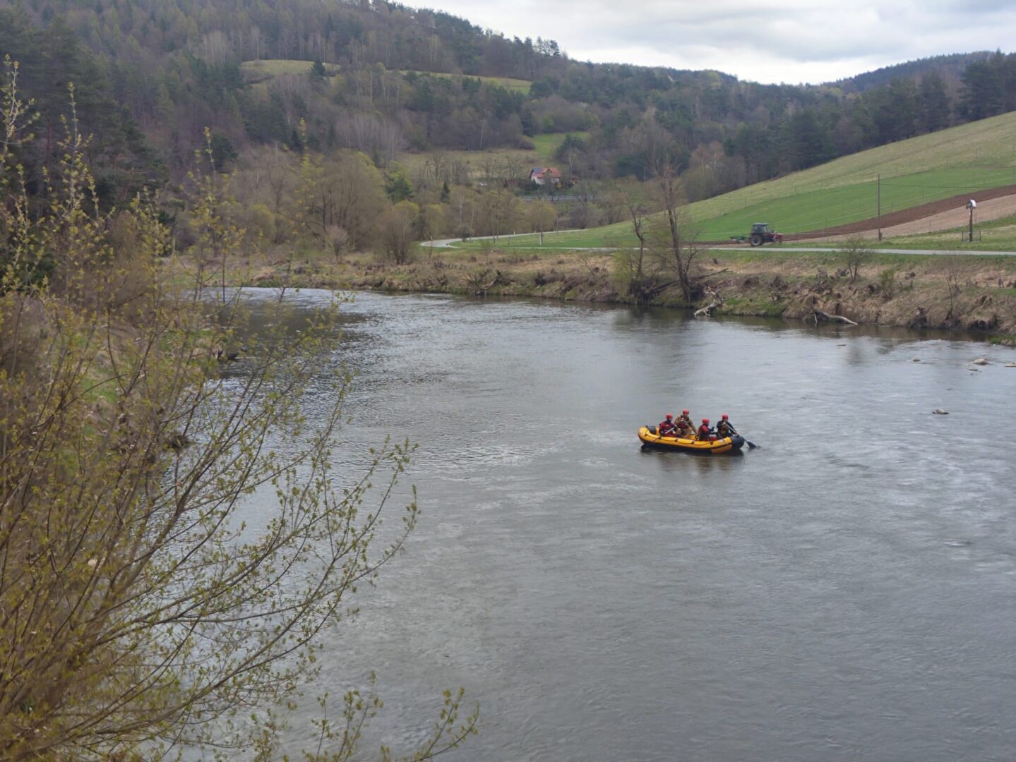 Six people in a yellow inflatable raft with red life jackets float on a wide river with green hills in the background and a tractor on a road nearby.