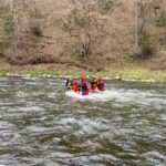 A group of people in red life jackets and helmets paddles together in a river near a wooded, rocky bank.