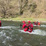 Group of rescuers in red wetsuits and helmets wading through a fast-flowing river during a training or rescue operation, with a wooded riverbank in the background.