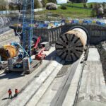 TBM tunnel construction site with a large circular tunnel boring machine, crane, and workers in orange vests inspecting the area
