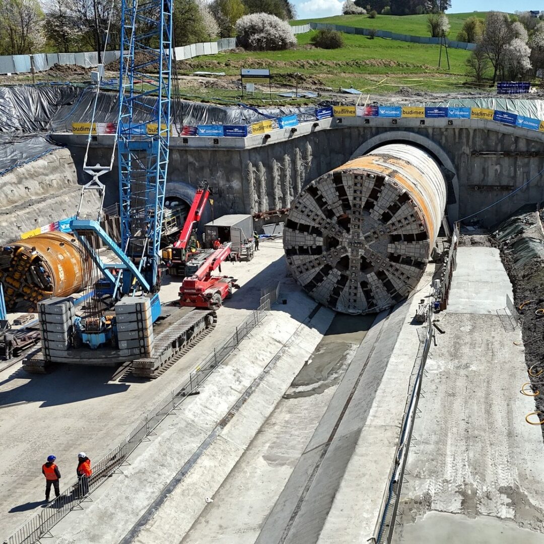 TBM tunnel construction site with a large circular tunnel boring machine, crane, and workers in orange vests inspecting the area