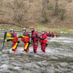 Group of rescuers in red-yellow dry suits and helmets crossing a river, paddling in a line toward the camera.
