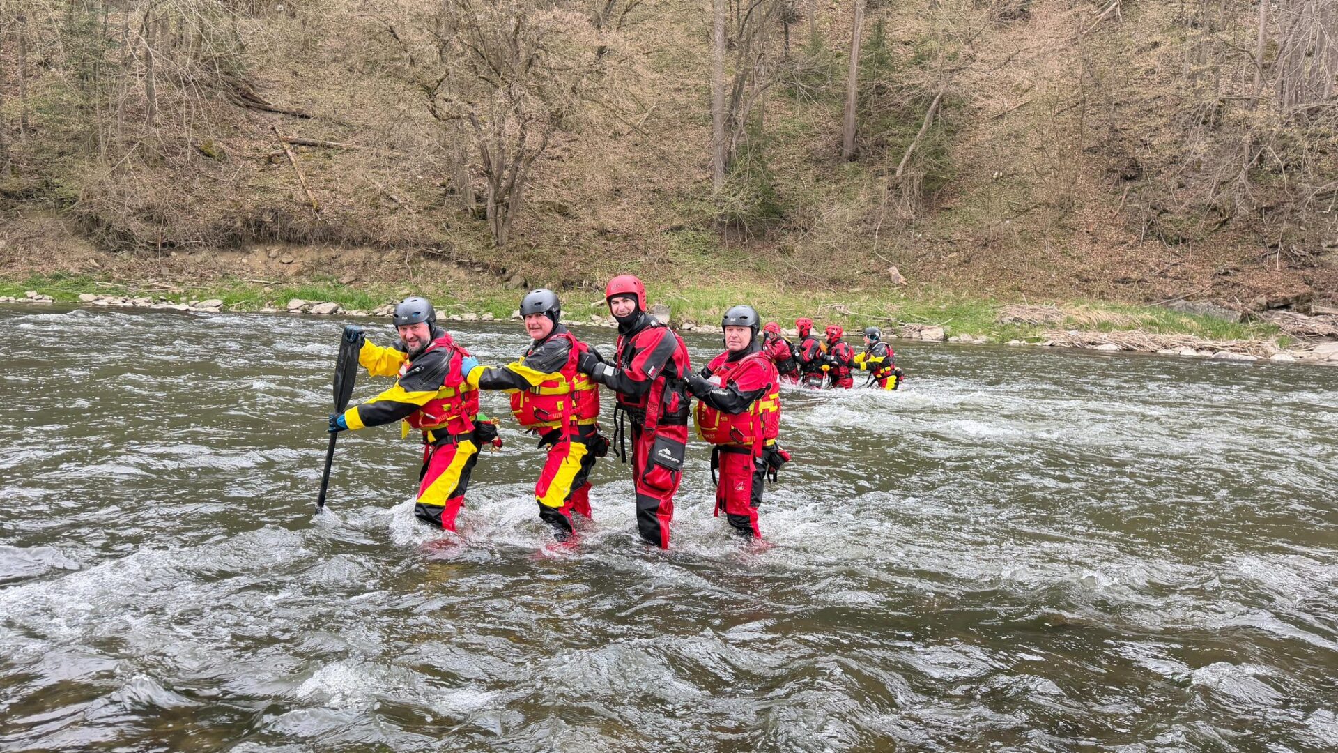 Group of rescuers in red-yellow dry suits and helmets crossing a river, paddling in a line toward the camera.