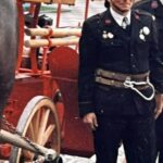 Firefighter in a vintage uniform with medals standing beside a red antique fire wagon on a cobblestone street.