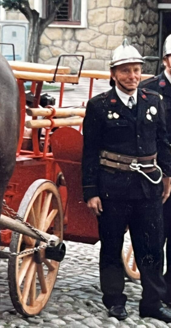 Firefighter in a vintage uniform with medals standing beside a red antique fire wagon on a cobblestone street.