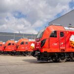 Red maintenance or passenger train locomotives lined up outside a large industrial building, showing a row of bright orange railcars with colorful mural art on the side.