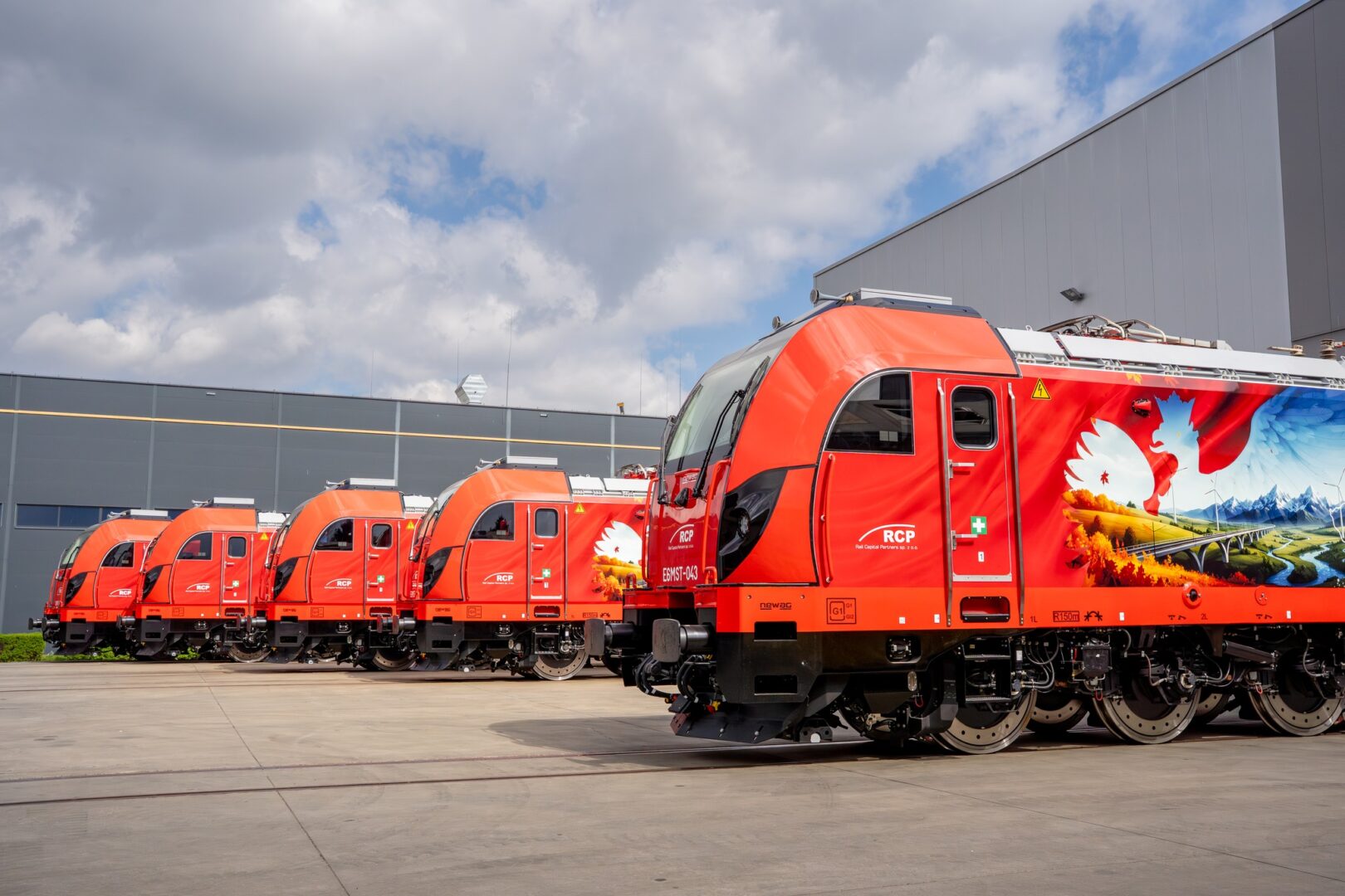 Red maintenance or passenger train locomotives lined up outside a large industrial building, showing a row of bright orange railcars with colorful mural art on the side.
