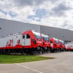 Row of red trains parked outside a large gray industrial building under a partly cloudy sky.