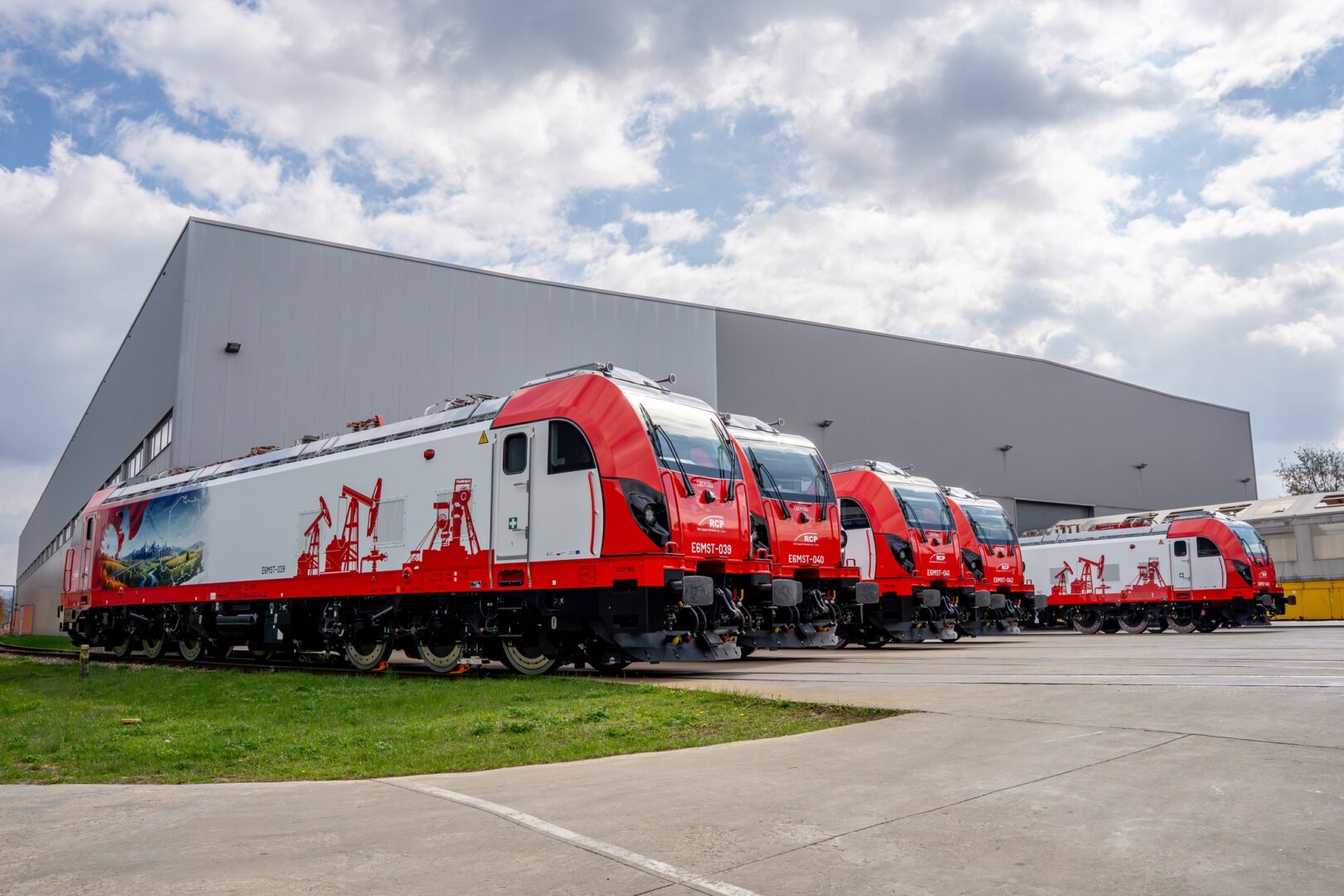 Row of red trains parked outside a large gray industrial building under a partly cloudy sky.
