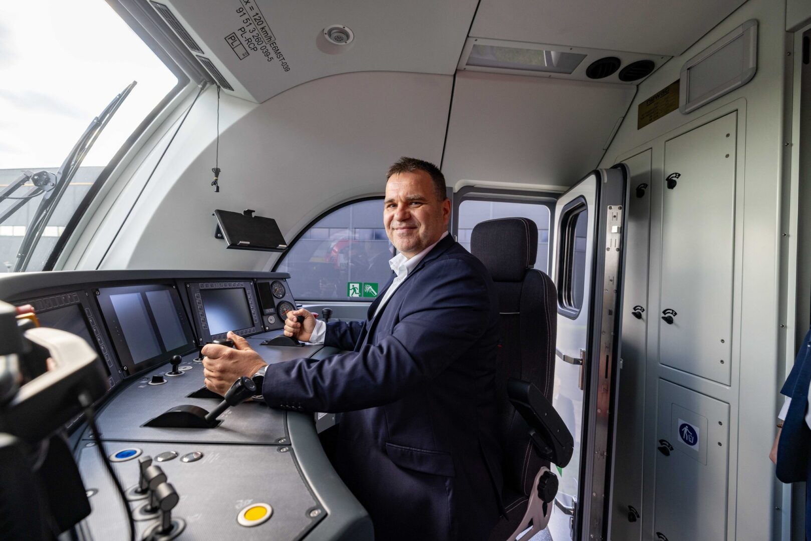 Male train operator in a navy suit sits at the cockpit controls, smiling, with instrument panel and large window behind him.
