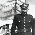 Black-and-white photo of a man in a vintage military uniform with a peaked cap, standing outdoors by a building.