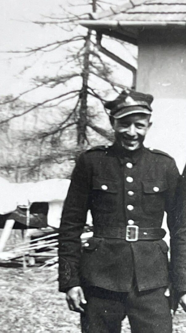 Black-and-white photo of a man in a vintage military uniform with a peaked cap, standing outdoors by a building.